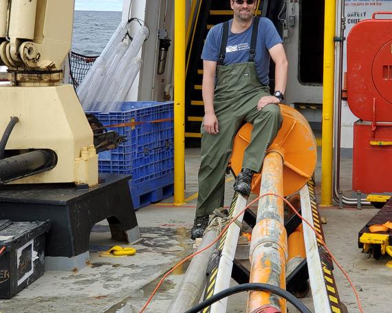 Guillaume St-Onge portant un casque orange de chantier, des lunettes de soleil et une salopette de pêcheur, assis au bout d’un carottier orange de plusieurs mètres de long.