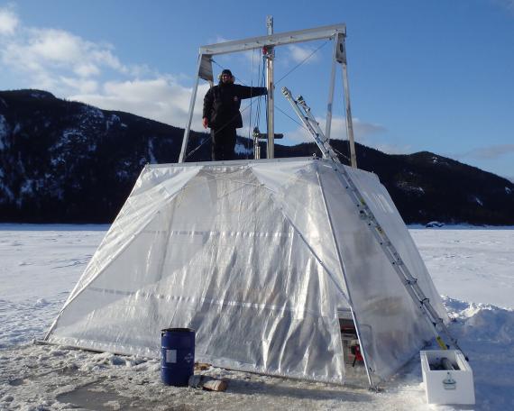 Guillaume St-Onge portant un manteau d’hiver et une tuque, posant au sommet d’un carottier protégé des intempéries par des bâches de plastique transparent sur un lac recouvert de neige.