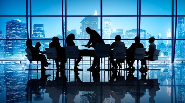 A stock photo of a black silhouette of people seated around a large boardroom table on a shiny, reflective floor and wall-to-wall windows in the background, showing a city skyline at night.