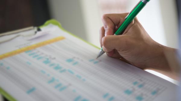 A stock photo of a blurred out clip board with a survey on it and a person's hand holding a green pen, filling out the survey.