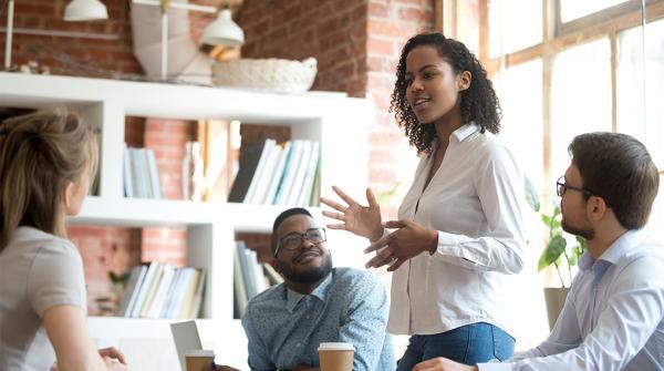 A stock image of men and women in dress shirts sitting around a table in a brick-walled office by a large window, looking up at a woman who is standing up at the table and talking to the group.