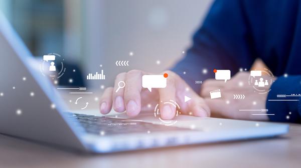 A stock image of a close up of someone's hands working at a laptop with white text message and other digital tech symbols floating around the hands.