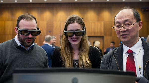 A photo of three people in a large conference room, looking at a computer screen, two wearing virtual reality goggles.